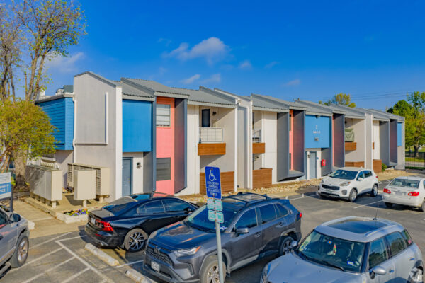 Row of modern townhomes with pastel blue, pink, and gray facades under a clear blue sky; cars parked in front.