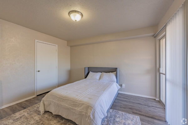 Small beige bedroom with a white bed and gray upholstered headboard, a white door on the left, and a large window with white vertical blinds on the right, illuminated by a ceiling light.