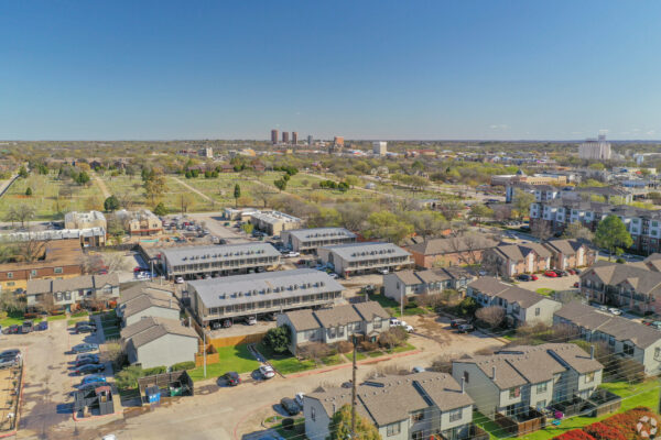 Aerial view of a suburban neighborhood with townhouses, parking lots, and a large park stretching toward the horizon under a clear blue sky.
