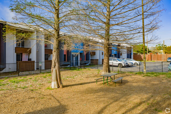 Leafless trees in a small outdoor courtyard in front of apartment buildings, with a black metal fence, a gravel yard, and a green picnic table area; several cars parked along the street beyond.