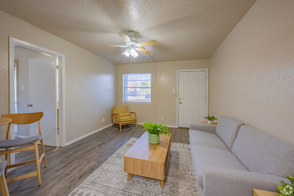 Living room interior with a gray sectional sofa, wooden coffee table, and ceiling fan; beige walls and a window with blinds in the background.