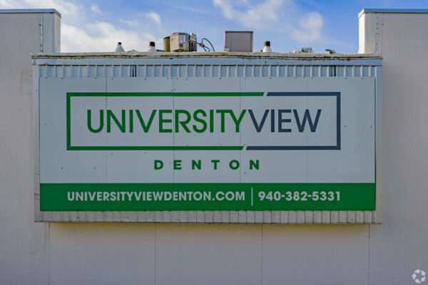 Signboard for 'University View Denton' mounted on an exterior wall, with green and blue framing against a blue sky.